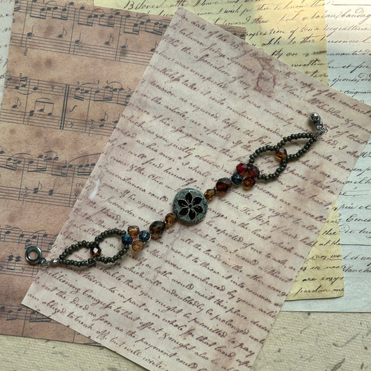 Antique Flower Window Bracelet with central floral medallion, amber and garnet Czech glass beads, bronze accents, flat-lay on vintage script paper