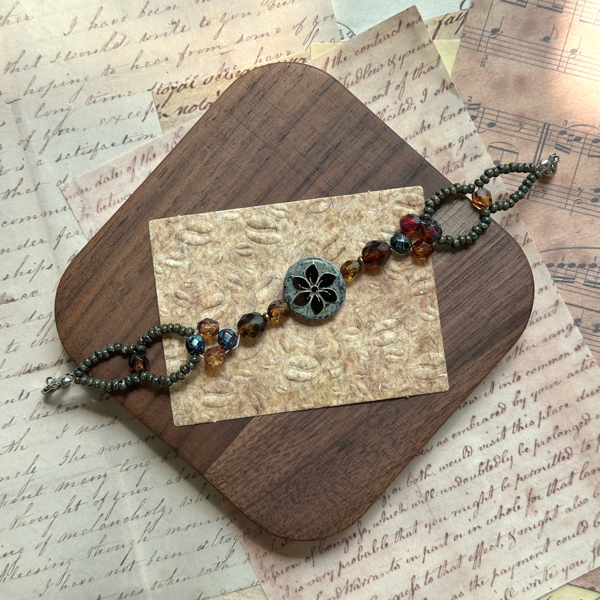 Artisan bracelet styled on a wooden board, floral medallion centerpiece with amber, garnet, and bronze glass beads, elegant oval seed-bead loops