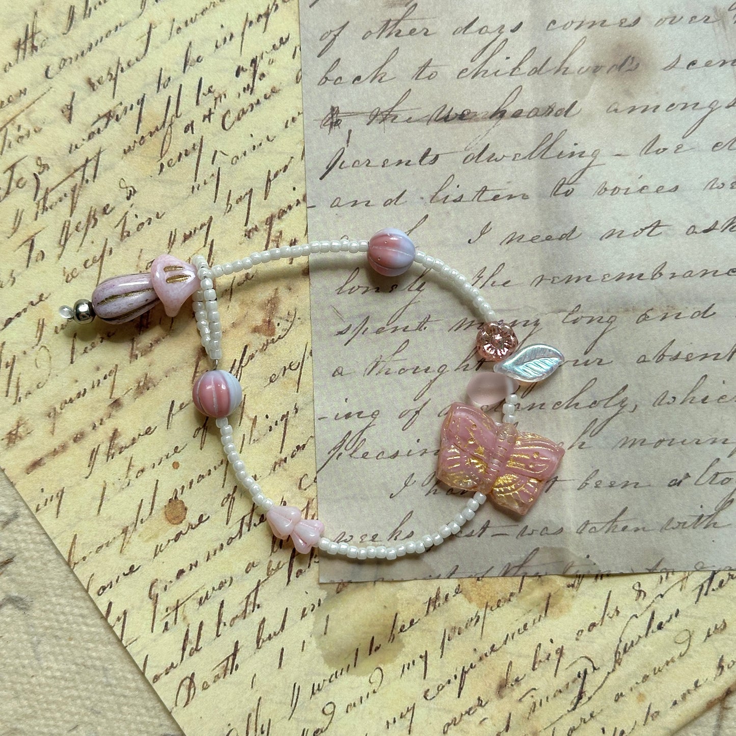 Blush Butterfly Dream Bracelet flat-lay on vintage script paper, pink butterfly bead with golden accents, paired with floral and pearly white glass beads