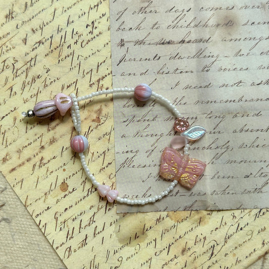 Blush Butterfly Dream Bracelet flat-lay on vintage script paper, pink butterfly bead with golden accents, paired with floral and pearly white glass beads