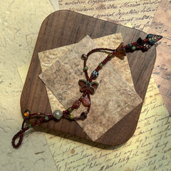 Artisan bracelet styled on a wooden board, deep jewel-tone beads with golden shimmer and leaf details around butterfly focal