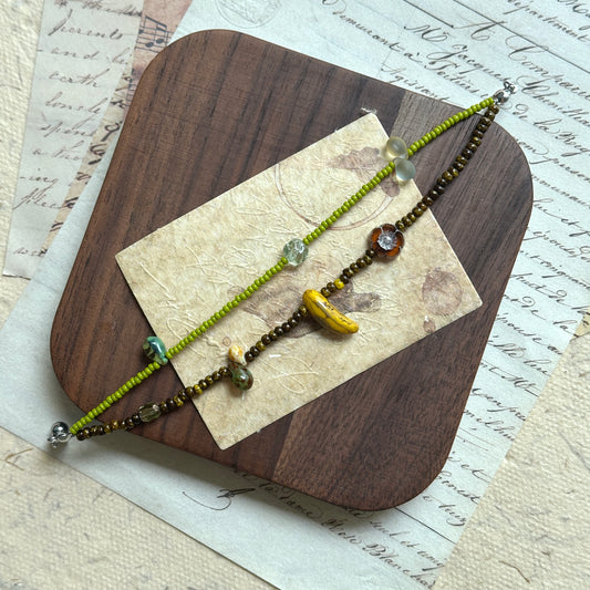 Orchard Twins Bracelet Set flat-lay on wooden board, lime-green beaded strand and bracelet with whimsical banana charm and amber accents
