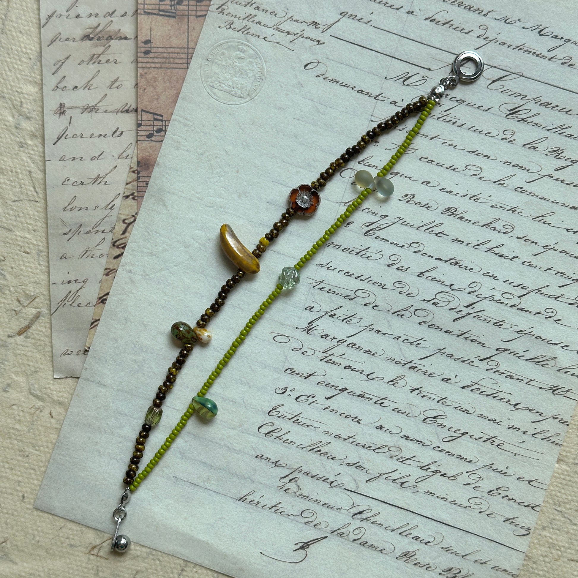 Close view of lime-green beaded bracelet on vintage script paper, glass charms and gold details in a fresh orchard palette