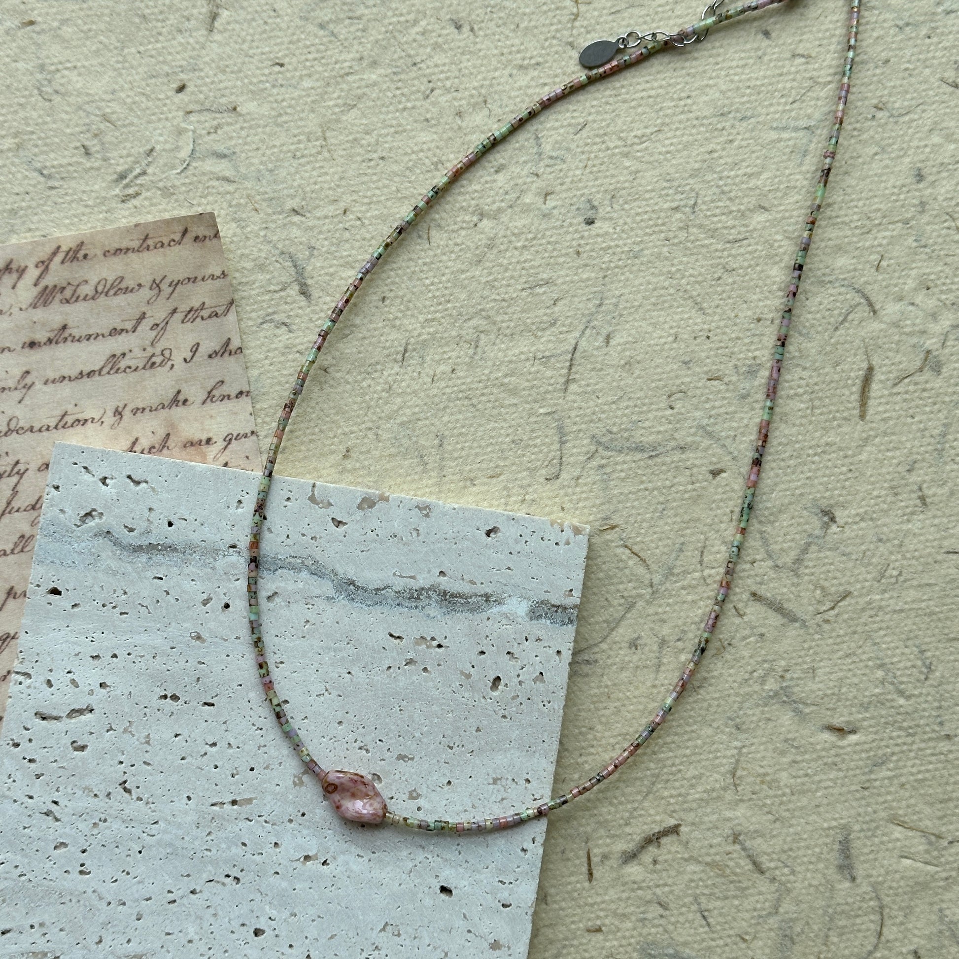 Morning Dew Petal Necklace flat-lay on vintage script paper, pastel seed beads in soft green and blush with a single pink floral focal