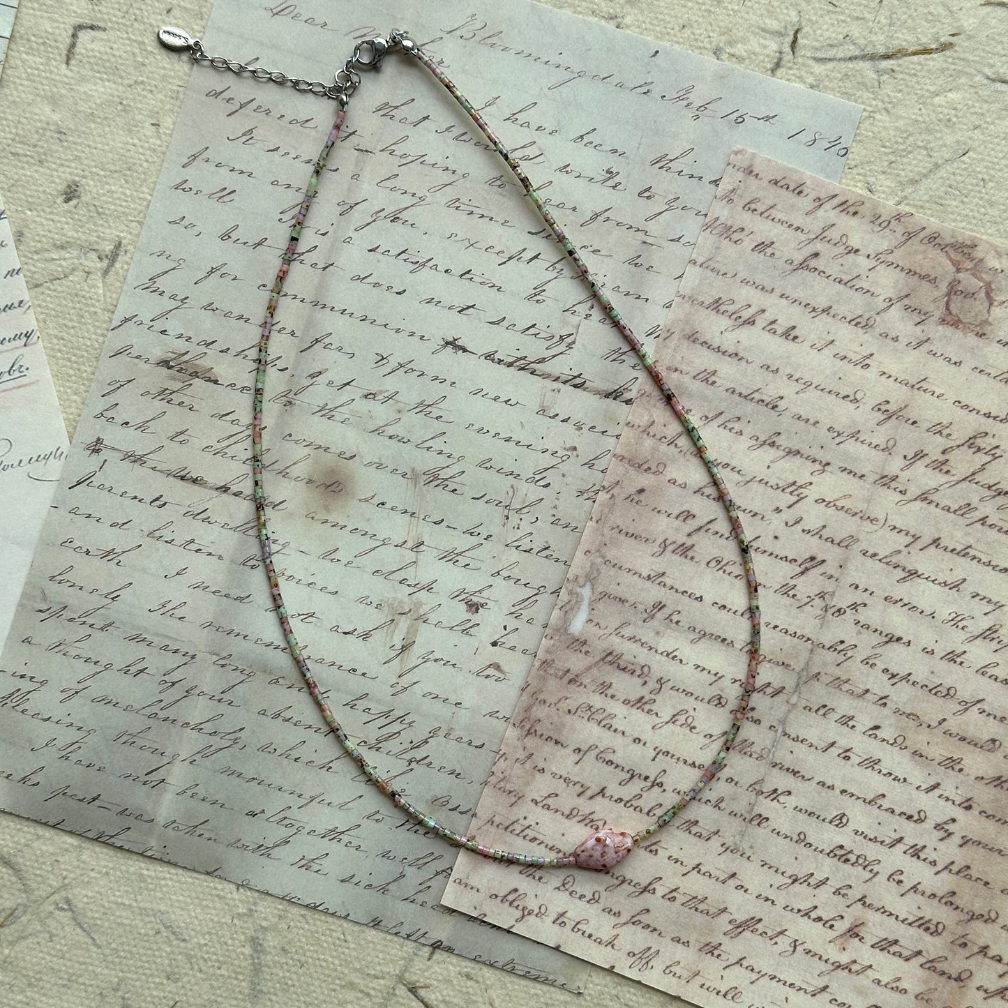 Delicate beaded necklace looped on paper background, minimalist strand of pastel seed beads with soft pink flower bead