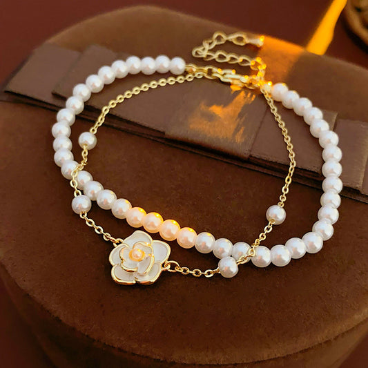 Layered faux-pearl bracelet with gold-tone chain and white enamel flower charm placed in a green jewelry tray — Petite Pearl Bloom Layered Bracelet.