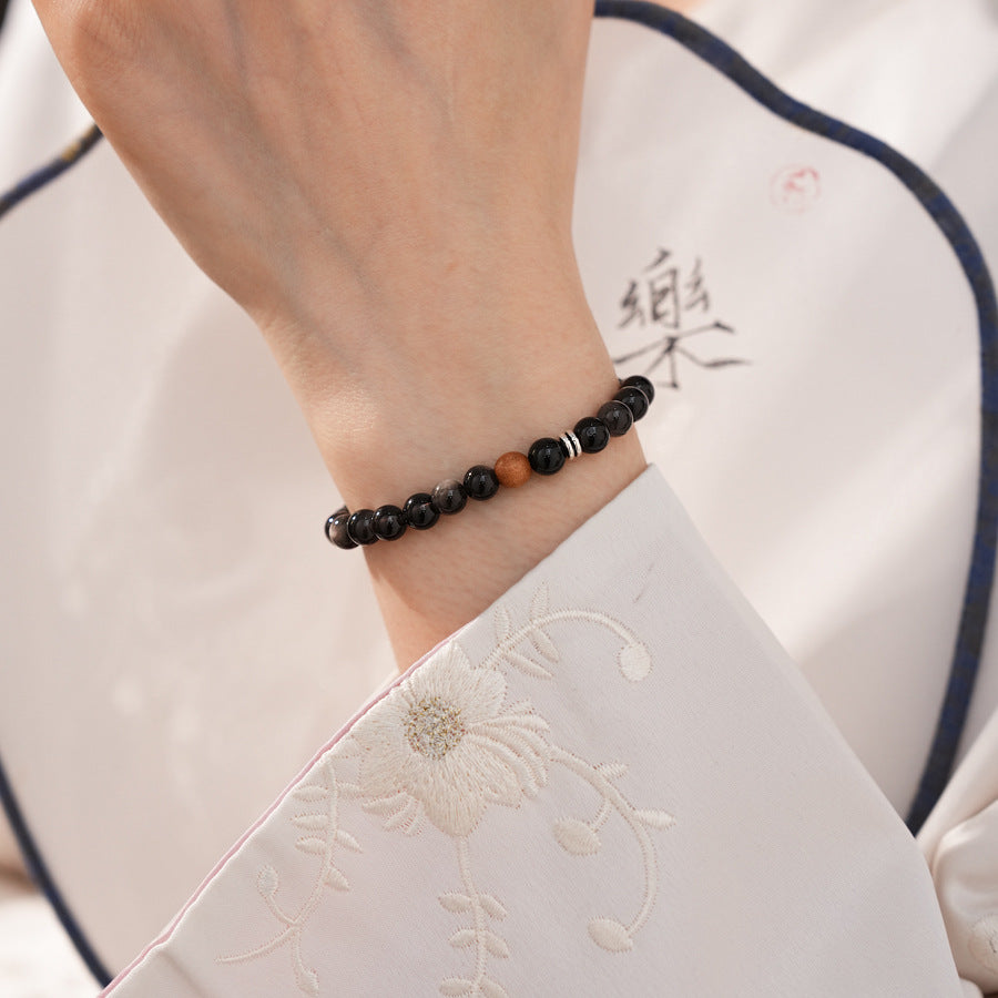 Sleek black silver-sheen stone beaded bracelet with one warm wood bead and silver ring spacers on a ceramic dish — Shadowwood Sheen Bracelet.