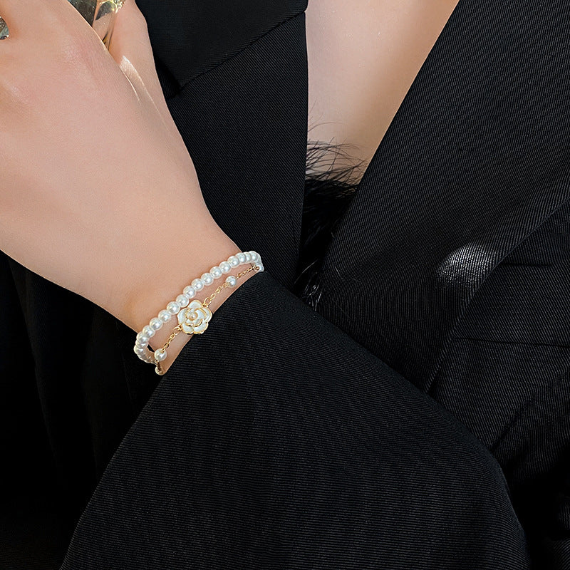 Layered faux-pearl bracelet with gold-tone chain and white enamel flower charm placed in a green jewelry tray — Petite Pearl Bloom Layered Bracelet.
