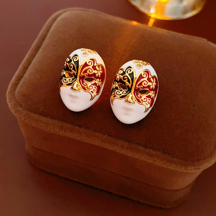 Enamel masquerade face stud earrings with red and black halves and gold filigree on a velvet jewelry box — Venetian Masquerade Studs.