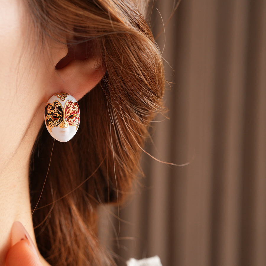 Enamel masquerade face stud earrings with red and black halves and gold filigree on a velvet jewelry box — Venetian Masquerade Studs.