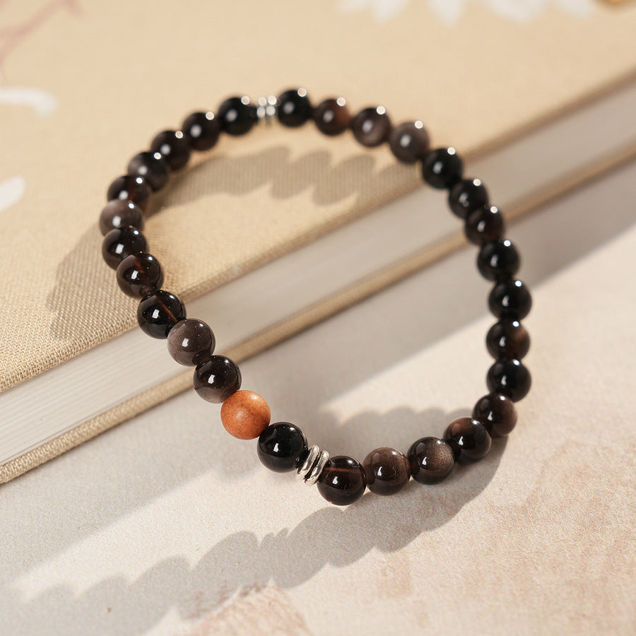 Sleek black silver-sheen stone beaded bracelet with one warm wood bead and silver ring spacers on a ceramic dish — Shadowwood Sheen Bracelet.