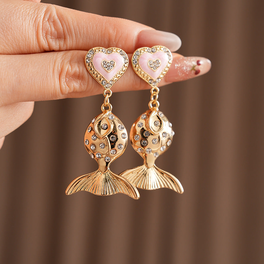 Gold-tone mermaid tail drop earrings with pink enamel heart studs and rhinestone accents on a green velvet background