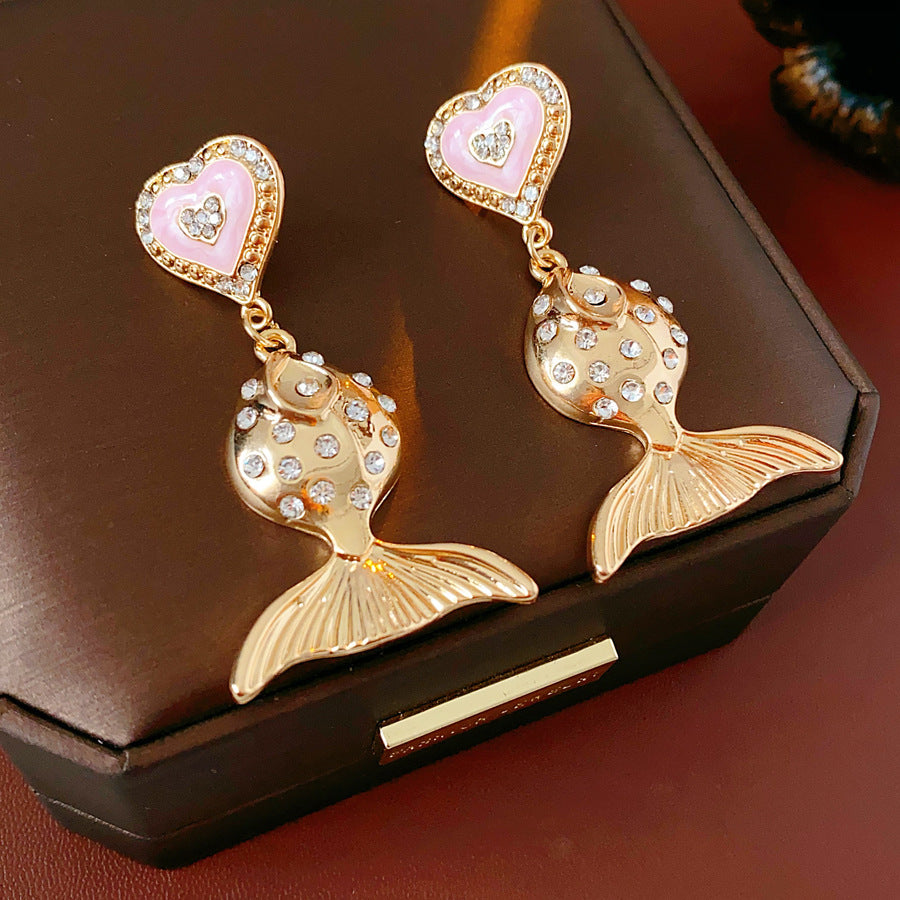 Gold-tone mermaid tail drop earrings with pink enamel heart studs and rhinestone accents on a green velvet background