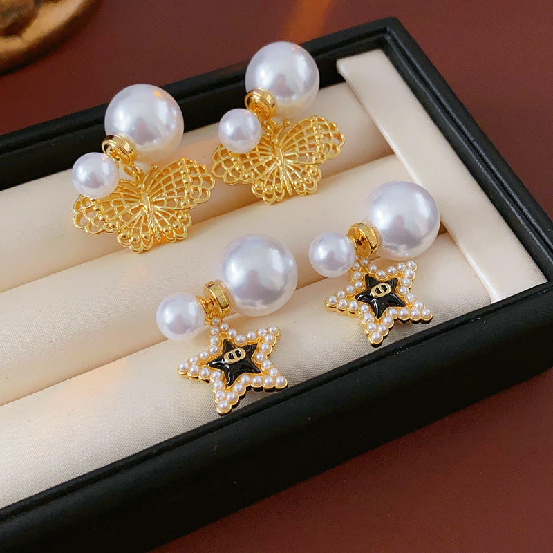 Double-pearl stud earrings with gold-tone butterfly and star charms displayed in a jewelry tray — Double Pearl Charm Studs.
