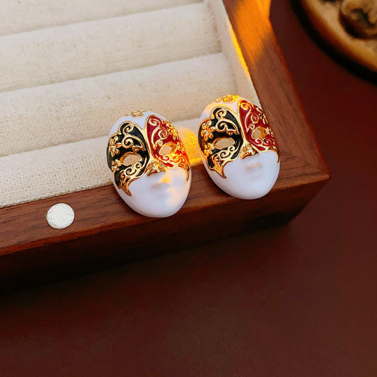 Enamel masquerade face stud earrings with red and black halves and gold filigree on a velvet jewelry box — Venetian Masquerade Studs.