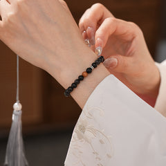Sleek black silver-sheen stone beaded bracelet with one warm wood bead and silver ring spacers on a ceramic dish — Shadowwood Sheen Bracelet.