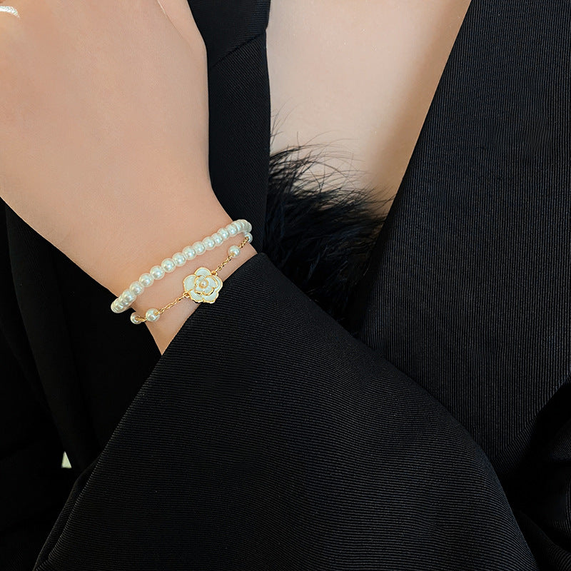 Layered faux-pearl bracelet with gold-tone chain and white enamel flower charm placed in a green jewelry tray — Petite Pearl Bloom Layered Bracelet.