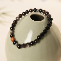 Sleek black silver-sheen stone beaded bracelet with one warm wood bead and silver ring spacers on a ceramic dish — Shadowwood Sheen Bracelet.