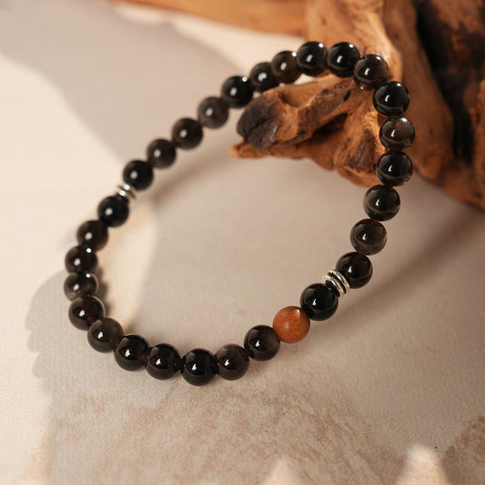 Sleek black silver-sheen stone beaded bracelet with one warm wood bead and silver ring spacers on a ceramic dish — Shadowwood Sheen Bracelet.