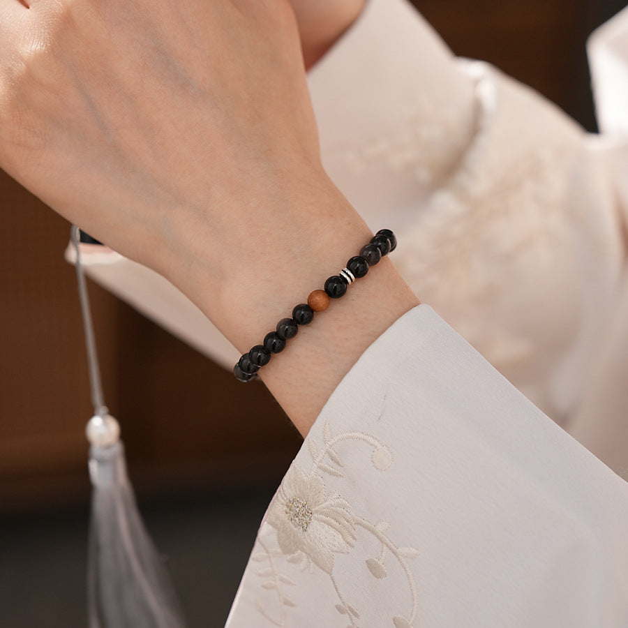 Sleek black silver-sheen stone beaded bracelet with one warm wood bead and silver ring spacers on a ceramic dish — Shadowwood Sheen Bracelet.