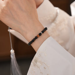 Sleek black silver-sheen stone beaded bracelet with one warm wood bead and silver ring spacers on a ceramic dish — Shadowwood Sheen Bracelet.