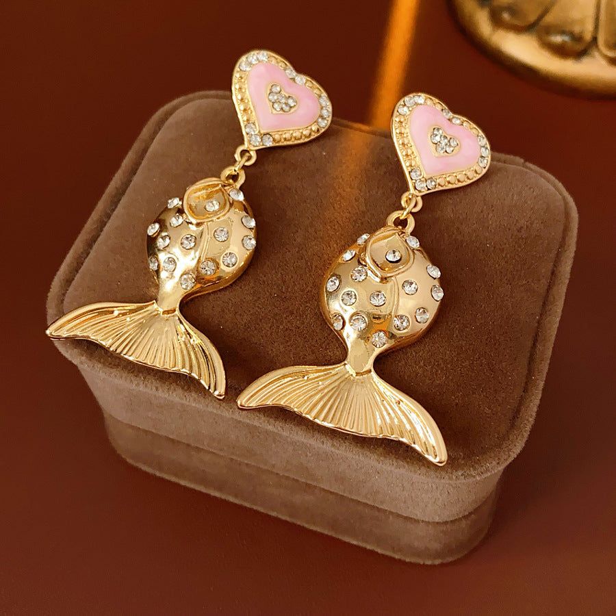 Gold-tone mermaid tail drop earrings with pink enamel heart studs and rhinestone accents on a green velvet background